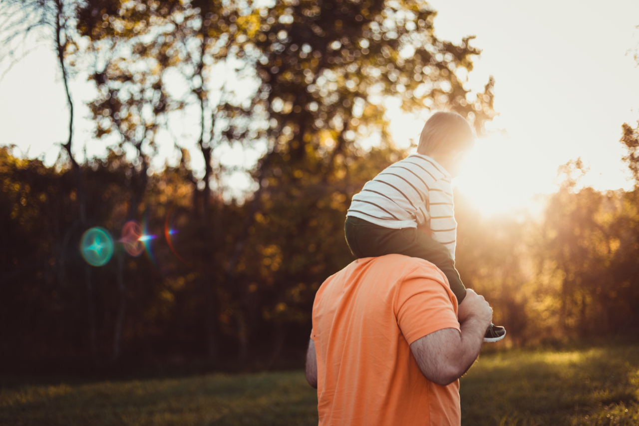 Man with son on shoulders walking away towards sunlight.